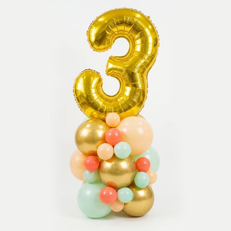 A birthday balloon decoration featuring a large metallic gold foil balloon shaped like the number 3, standing on top of a cluster of assorted balloons in gold, peach, coral, mint green, and light blue, set against a plain white background.