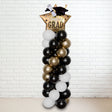 A tall balloon column with a graduation theme, featuring black, gold, and white balloons, topped with a 'GRAD' hat.