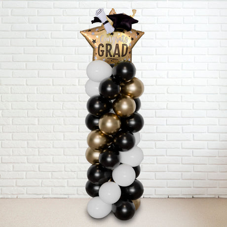 A tall balloon column with a graduation theme, featuring black, gold, and white balloons, topped with a 'GRAD' hat.