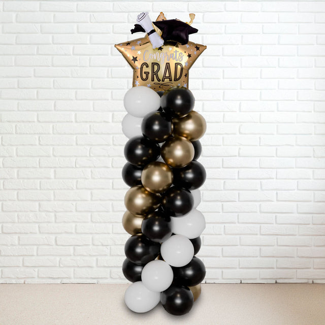 A tall balloon column with a graduation theme, featuring black, gold, and white balloons, topped with a 'GRAD' hat.