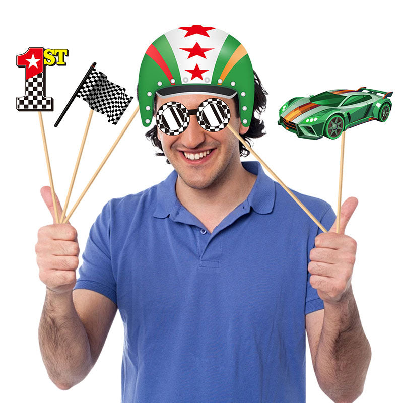 A man wearing a green, white, and red racing helmet with striped novelty glasses holds up race-themed props on sticks, including a first-place finish line, a checkered flag, and a green race car, against a plain white background.