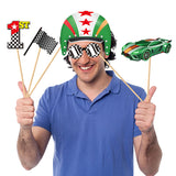 A man wearing a green, white, and red racing helmet with striped novelty glasses holds up race-themed props on sticks, including a first-place finish line, a checkered flag, and a green race car, against a plain white background.